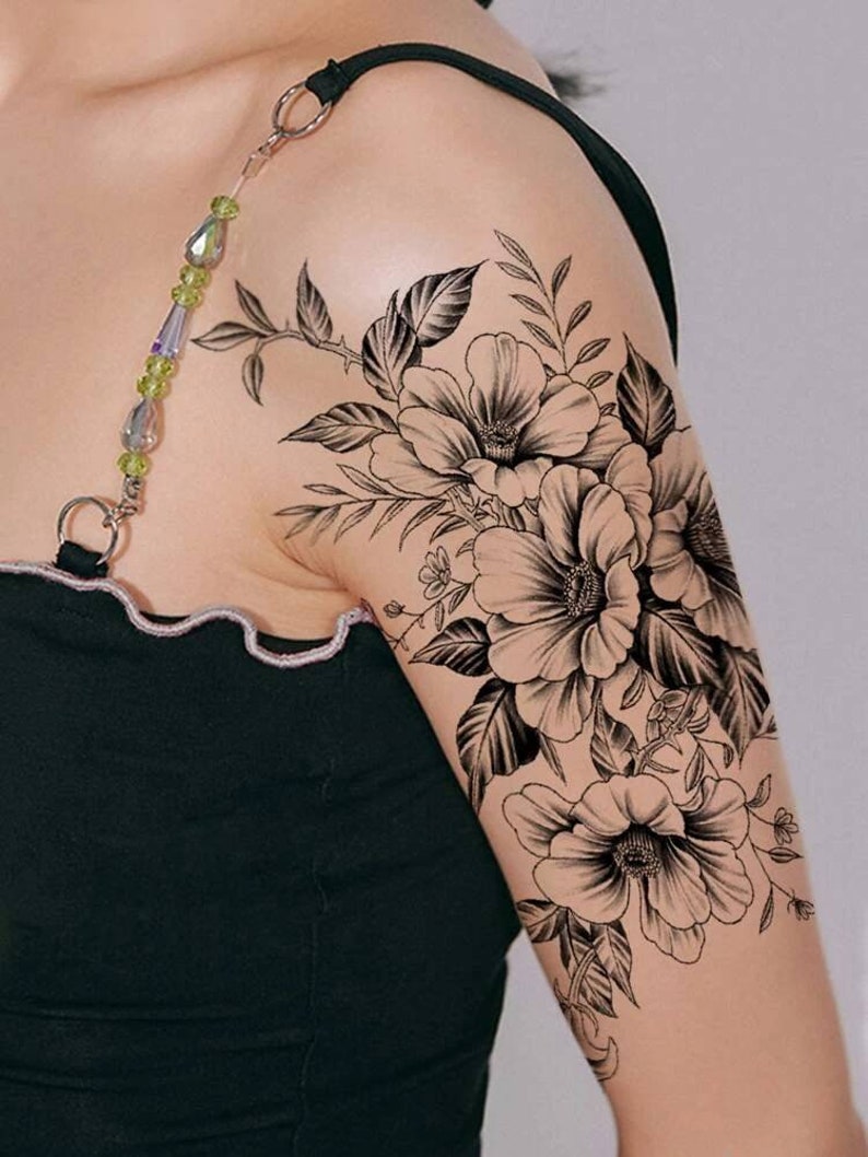 May include: Black and white floral tattoo design featuring three large flowers with detailed petals and leaves. The tattoo is on a person's upper arm.