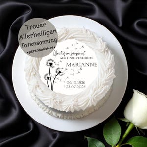 Könnte beinhalten: Ein weißer Kuchen mit dekorativem Rand, belegt mit einem kreisförmigen essbaren Bild. Das Bild zeigt Pusteblumen-Illustrationen und den Text "Was tief im Herzen ist, GEHT NIE VERLOREN. MARIANNE 06.10.1936 + 23.07.2025". Eine weiße Rose ist in der Ecke.