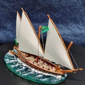 Op de afbeelding: Gedetailleerd model zeilboot met witte zeilen en een bruine houten romp. Het miniatuurschip heeft twee masten en staat op een basis die oceaangolven uitbeeldt. Een groene vlag is zichtbaar op de mast. Het model is een gedetailleerde weergave.