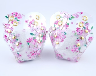 May include: A pair of clear, heart-shaped face shields. The shields are decorated with pink cherry blossom branches and gold-colored eyelets. The interior features a white fabric lining.