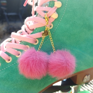 Può includere: Un paio di pompon rosa e soffici attaccati a una catena dorata con un ciondolo a forma di cuore. La catena è attaccata a un pattino a rotelle verde.