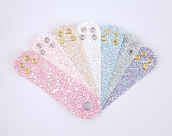 May include: A collection of seven glittery leatherette wrist cuffs in a fan arrangement. Each cuff is a different pastel color, including pink, gold, and blue. They have eyelets with gold and silver grommets and a single hole at the end.