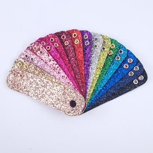 Könnte beinhalten: Ein Fächer aus glitzernden Kunstleder-Mustern in verschiedenen Farben, darunter Gold, Rosa, Rot, Lila, Grün und Blau. Jedes Muster hat eine Metallöse und ist an einem zentralen schwarzen Stück befestigt.