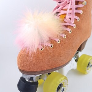 Pu&ograve; includere: Un paio di pattini a rotelle in camoscio marrone con lacci rosa e un pompon rosa e bianco soffice sulla punta.