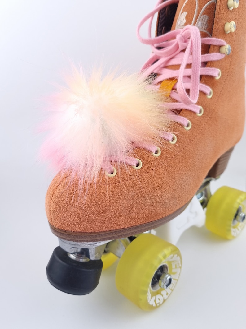 Pu&ograve; includere: Un paio di pattini a rotelle in camoscio marrone con lacci rosa e un pompon rosa e bianco soffice sulla punta.