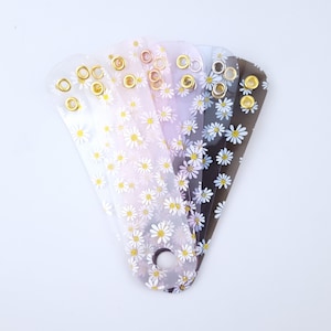 May include: Five clear plastic accessories with a daisy pattern. Each has a different colour gradient, from clear to black, and gold-coloured eyelets. The daisy design features white petals and yellow centres.