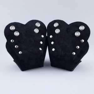 Könnte beinhalten: Zwei schwarze herzförmige Accessoires mit silbernen Ösen und Nieten. Die Artikel haben eine weiche, Wildleder-ähnliche Textur und ein geschichtetes Design. Das symmetrische Design zeigt jedes Teil spiegelbildlich, mit einem dunklen, samtigen Aussehen.