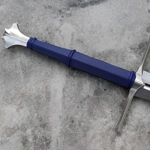 May include: A longsword featuring a polished silver-colored metal blade, a crossguard, and a deep blue handle with a silver-colored pommel. A matching blue scabbard is positioned alongside the sword, showing a curved opening at the top.