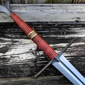 May include: A longsword featuring a double-edged steel blade, a crossguard, and a grip wrapped in reddish-brown leather with gold-coloured wire accents. The hilt includes a central brass-toned ring and a flared, metallic pommel at the end.