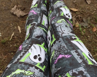 May include: Leggings featuring a repeating pattern of white ghosts, purple and lime green bats, and black spiderwebs on a dark gray background. The design includes rectangular frames and scattered stars, creating a festive Halloween-themed graphic print.