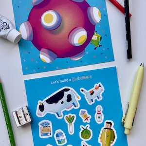 May include: Two illustrated cards with a space theme and the text "Let's build a Katamari". The cards are surrounded by art supplies, including pencils, erasers, and paint tubes. The top card features a pink planet with white and yellow accents.