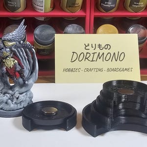 May include: A set of black plastic, tiered, magnetic display stands. The stands are shaped like a crescent moon and are designed to hold miniature figures. The stands are on a table next to a miniature figure of a winged warrior in silver armor. The text "どりもの DORIMONO HOBBIES-CRAFTING-BOARDGAMES" is on a yellow note card.