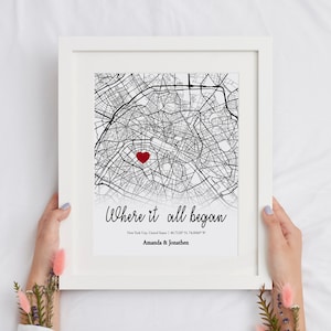 Op de afbeelding: Een zwart-witte kaartprint met een rood hart dat een locatie markeert. De tekst "Where it all began" is in cursief boven de kaart geschreven. Onder de kaart staat de tekst "New York City, United States | 40.7128&deg; N, 74.0060&deg; W" en "Amanda & Jonathen".