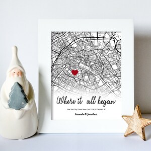 Op de afbeelding: Een ingelijste zwart-witte kaart van New York City met een rood hart dat een locatie markeert. De tekst "Where it all began" staat onder de kaart, samen met de co&ouml;rdinaten "40.7128&deg; N 74.0060&deg; W" en de namen "Amanda & Jonathen".