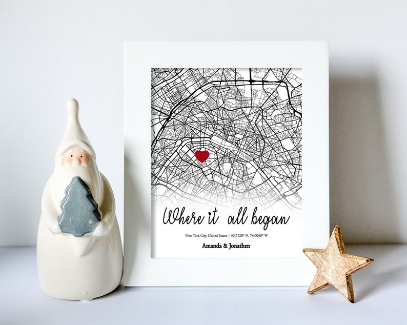 Op de afbeelding: Een ingelijste zwart-witte kaart van New York City met een rood hart dat een locatie markeert. De tekst "Where it all began" staat onder de kaart, samen met de co&ouml;rdinaten "40.7128&deg; N 74.0060&deg; W" en de namen "Amanda & Jonathen".
