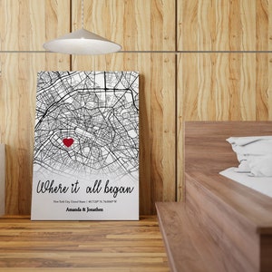 Op de afbeelding: Een zwart-witte kaartprint met een rood hart dat een locatie markeert. De tekst "Where it all began" staat onder de kaart, samen met de stad, de staat en de co&ouml;rdinaten. De namen "Amanda & Jonathan" staan onder de co&ouml;rdinaten.