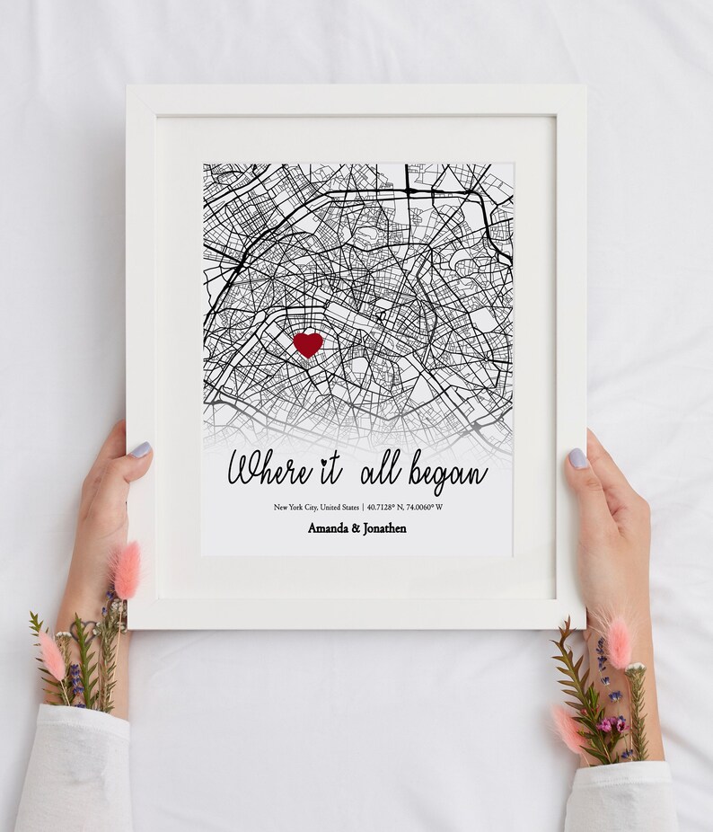 Op de afbeelding: Een zwart-witte kaartprint met een rood hart dat een locatie markeert. De tekst "Where it all began" is in cursief boven de kaart geschreven. Onder de kaart staat de tekst "New York City, United States | 40.7128&deg; N, 74.0060&deg; W" en "Amanda & Jonathen".