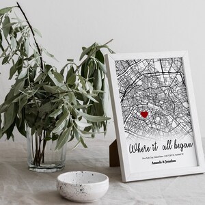 Op de afbeelding: Een zwart-witte kaartprint met een rood hart dat een locatie markeert. De print is wit ingelijst en heeft de tekst "Where it all began" met de stad, staat en co&ouml;rdinaten eronder. De namen "Amanda & Jonathan" staan onderaan de print.