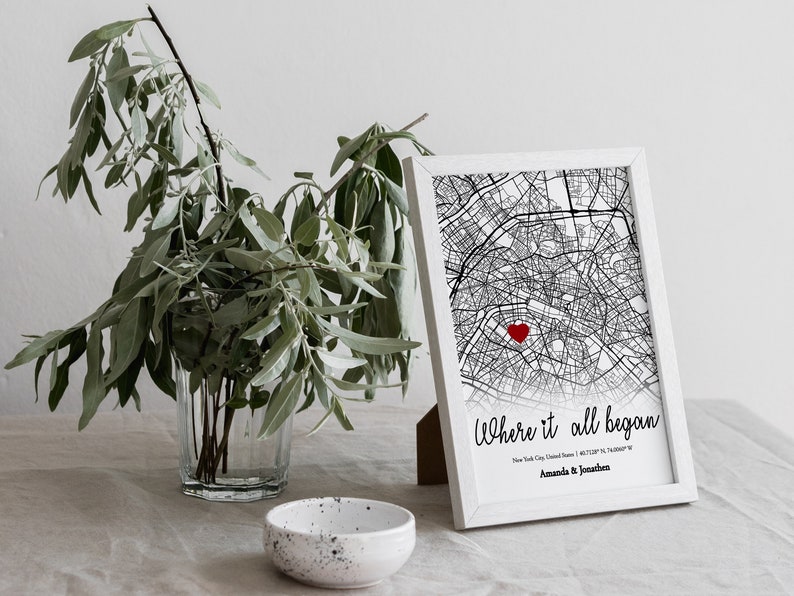 Op de afbeelding: Een zwart-witte kaartprint met een rood hart dat een locatie markeert. De print is wit ingelijst en heeft de tekst "Where it all began" met de stad, staat en co&ouml;rdinaten eronder. De namen "Amanda & Jonathan" staan onderaan de print.