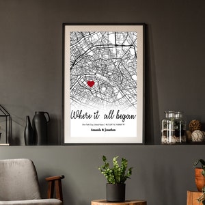 Op de afbeelding: Zwart-witte stadskaartprint met een rood hart dat een locatie markeert. De tekst "Where it all began" staat onder de kaart met de stad, de staat en de co&ouml;rdinaten. De namen "Amanda & Jonathan" staan onder de co&ouml;rdinaten.