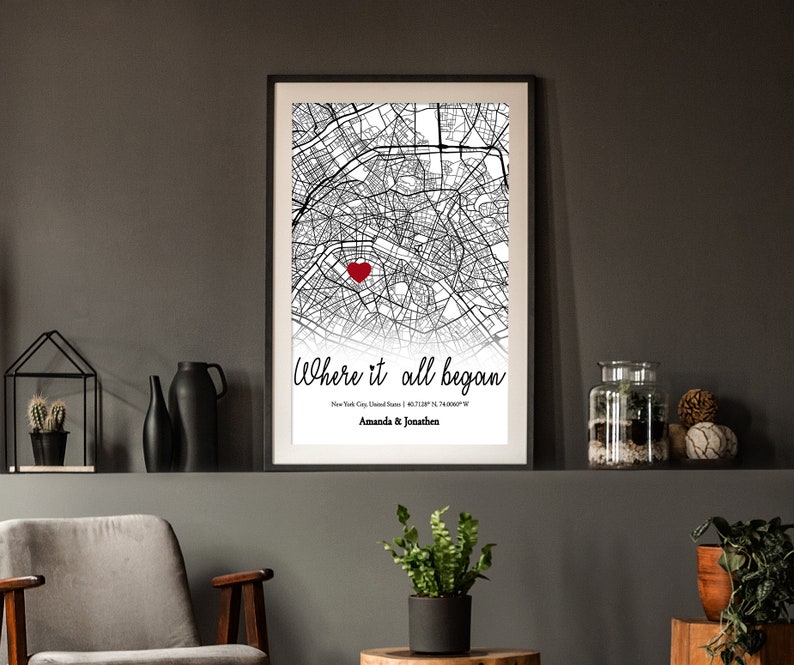 Op de afbeelding: Zwart-witte stadskaartprint met een rood hart dat een locatie markeert. De tekst "Where it all began" staat onder de kaart met de stad, de staat en de co&ouml;rdinaten. De namen "Amanda & Jonathan" staan onder de co&ouml;rdinaten.