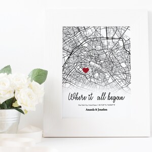 Op de afbeelding: Een zwart-witte kaartprint met een rood hart dat de locatie van New York City, Verenigde Staten, markeert. De tekst "Where it all began" staat onder de kaart, gevolgd door de co&ouml;rdinaten en de namen "Amanda & Jonathan".