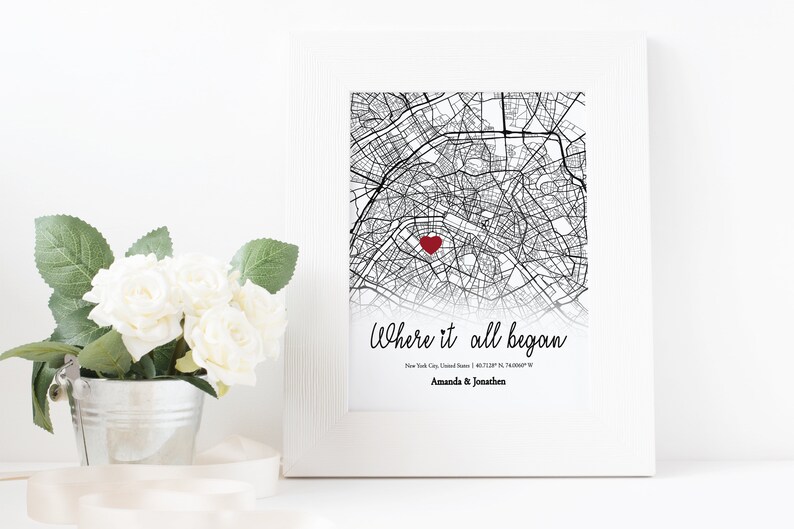 Op de afbeelding: Een zwart-witte kaartprint met een rood hart dat de locatie van New York City, Verenigde Staten, markeert. De tekst "Where it all began" staat onder de kaart, gevolgd door de co&ouml;rdinaten en de namen "Amanda & Jonathan".