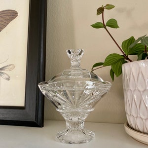 May include: A clear glass candy dish with a lid and a pedestal base. The dish has a faceted design and is decorated with a geometric pattern.