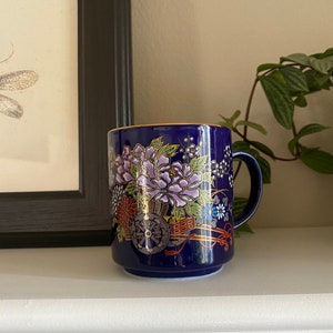 May include: A blue ceramic mug with a gold rim and a floral design. The design features a cart with a wheel, flowers, and leaves in gold, red, and white.