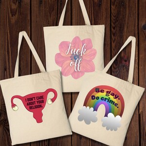 Könnte beinhalten: Drei beigefarbene Canvas-Tragetaschen mit verschiedenen grafischen Designs. Eine zeigt eine rosa Blume mit den Worten "Fuck off". Eine andere zeigt eine Gebärmutter mit dem Text "I don't care about your religion." Die dritte Tasche hat einen Regenbogen und die Worte "Be gay. Do crime."