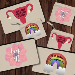 May include: A collection of beige canvas zipper pouches with various designs. Some pouches feature a uterus graphic with the text "I DON'T CARE ABOUT YOUR RELIGION." Others display a rainbow with "Be gay. Do crime." and a pink flower with "Fuck off."
