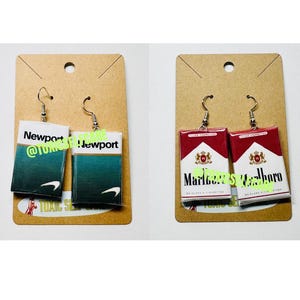Op de afbeelding: Oorbellen met miniatuurreplica's van sigarettenpakjes. Eén paar toont een Newport-pakje in groen en wit, het andere een Marlboro-pakje in rood en wit. Beide paren hebben zilverkleurige oorhaakjes.