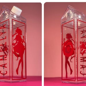 May include: Clear plastic milk carton-shaped bottle with a white cap. The bottle has red text and a red silhouette of a woman. The text reads "CAMELIA".