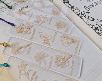 May include: Set of ten clear acrylic bookmarks with gold floral designs and names of flowers. The bookmarks are arranged in a fan shape on a white surface. The text on the bookmarks reads: "Water Hyacinth", "Phlox", "Queen Ann's Lace", "Glory Lily", "Iris Germanica", "Six O'Clock", "Woodrow Aster", "Cosmos", "Sweet Pea", "Gelsemium".