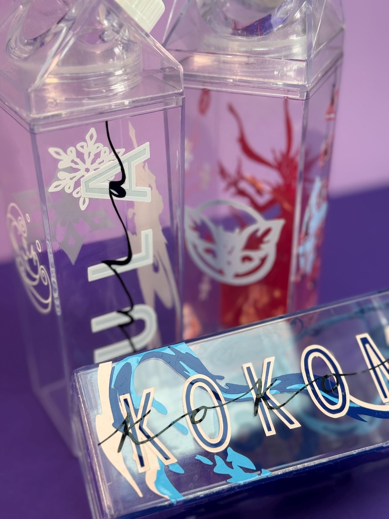 May include: Clear plastic water bottles with decorative designs. One bottle features a snowflake and the word "N", another has a red tree design, and a third has the word "KOKON" in blue and white. The bottles have a milk carton shape.