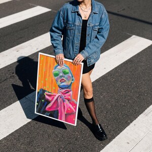 Op de afbeelding: Een ingelijst portret met een gestileerde illustratie van een persoon met een blauwe huid, groene zonnebril en een roze strik. De achtergrond is oranje met groene stippen. De lijst is wit.