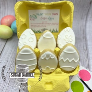 May include: A yellow egg carton holds six decorated Easter egg cookies with white icing designs. The carton includes a card that says "Paint Your Own Easter Eggs". A small paint palette and brush are also included.