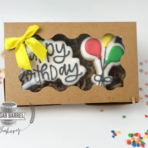 May include: A brown cardboard box with a window showing two decorated sugar cookies. One cookie says "Happy Birthday" and the other has three balloons. The box is tied with a yellow ribbon.