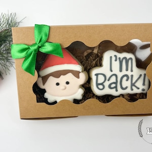 May include: A brown box with a green bow on top. The box contains two decorated sugar cookies. One cookie is an elf's face and the other cookie says "I'm Back!" The box is labeled "Sugar Barrel Bakery".