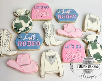 Może przedstawiać: Asortyment zdobionych ciasteczek cukrowych o tematyce rodeo. Ciasteczka zawierają kowbojskie buty, kapelusze i kurtki z napisami "Let's Go Girls" i "Last Rodeo" w różowym i niebieskim lukrze. Niektóre ciasteczka mają srebrne akcenty.