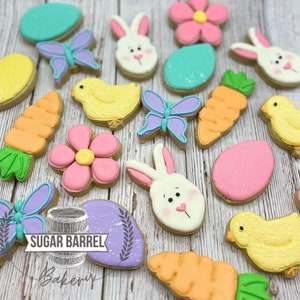 May include: A collection of Easter-themed sugar cookies decorated with various designs, including bunnies, chicks, butterflies, carrots, flowers, and eggs. The cookies are arranged on a light wood background. The text "Sugar Barrel Bakery" is visible on one of the cookies.
