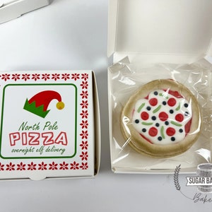 May include: A white pizza box with a red and green border and a festive elf design. The box says "North Pole Pizza overnight elf delivery". A white pizza cookie with red, green, and black toppings is in a clear plastic bag inside a white box with the words "Sugar Barrel Bakery" on the bottom.
