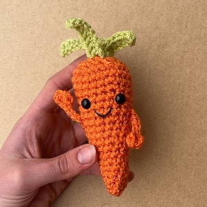 May include: A crocheted carrot toy with a smiling face and green leafy top. The carrot is orange and has black eyes and a mouth.