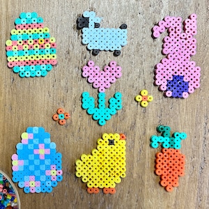 Könnte beinhalten: Ein Set aus bunten Perlen-Sprites zum Thema Ostern, darunter ein Küken, ein Hase, eine Karotte, ein Ei und Blumen. Die Sprites bestehen aus kleinen, bunten Perlen.