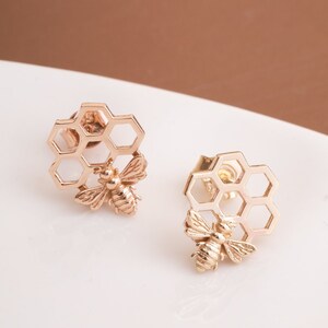 May include: A pair of gold-toned earrings featuring a bee design on a honeycomb background.