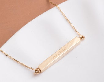 May include: A gold-colored necklace with a rectangular pendant. The pendant is engraved with the name "Archie" in a simple font. The necklace is displayed on a white surface, with a brown background.
