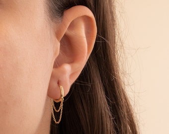 May include: A gold hoop earring with a delicate chain dangling from it. The earring is worn in the left ear of a person with long brown hair.