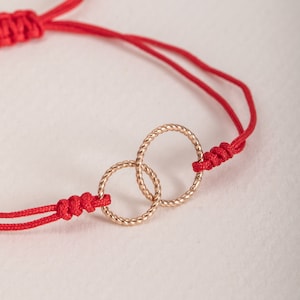 以下が含まれることがあります： 赤い紐のブレスレットに、2つの金色の輪が絡み合っています。輪はねじれていて、繊細なデザインです。