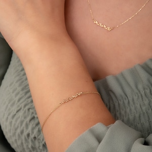 May include: A gold chain bracelet with the word "mama" spelled out in cursive. The bracelet is on a person's wrist.