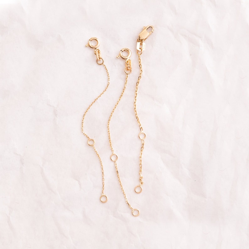 Three gold chain extenders of different lengths. Each extender has a lobster clasp on one end and a jump ring on the other end.